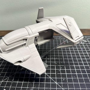 Peut inclure: Modèle imprimé en 3D, de couleur gris clair, représentant un vaisseau spatial futuriste. Le modèle présente un design détaillé avec des ailes, une zone de cockpit et divers composants angulaires. Sur une surface quadrillée noire et grise.