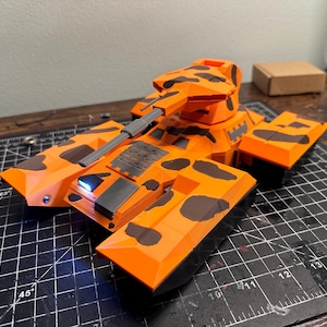 May include: A detailed orange and black toy tank model. The tank has a rotating turret, a long barrel, and side panels. The model features a camouflage pattern and is set on a cutting mat.