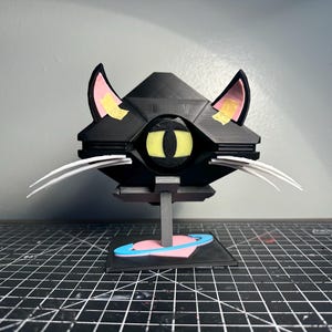 Op de afbeelding: Een zwart katvormig beeldhouwwerk met gele ogen, roze binnenoren en witte snorharen. Het beeldhouwwerk is gemonteerd op een standaard met een hart- en planeetontwerp aan de basis. De oren hebben gele tape.