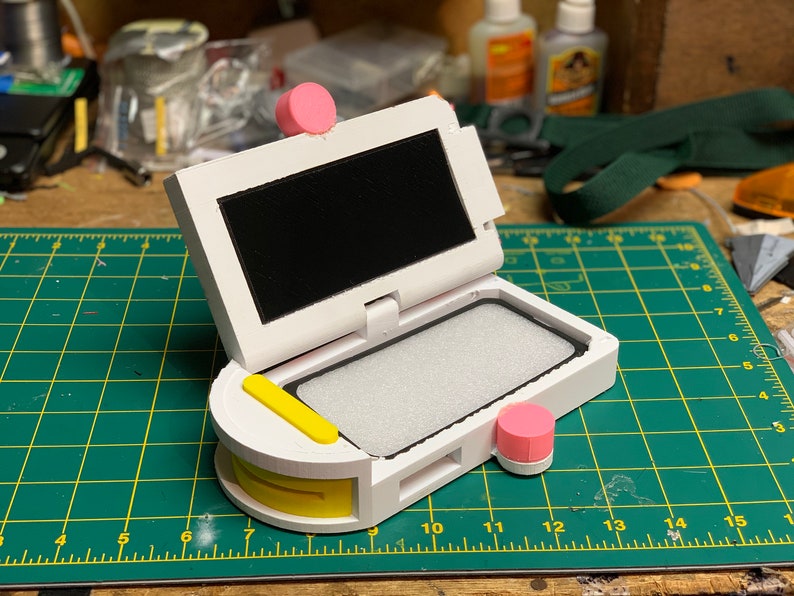 Puede incluir: Una caja impresa en 3D de color blanco y amarillo con una pantalla negra y botones rosas. La caja est&aacute; abierta y tiene un forro de espuma blanca en el interior.