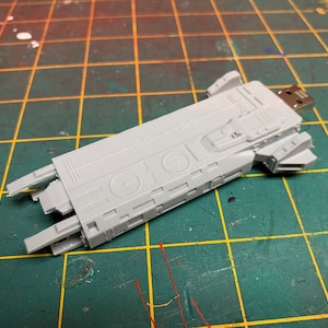 Puede incluir: Un modelo de nave espacial de plástico gris con un conector USB. El modelo está detallado con líneas y círculos en relieve.