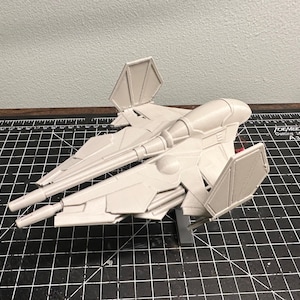 May include: A light gray model of a futuristic spacecraft, featuring a central fuselage, two forward-extending cannons, and angled wings. The model is resting on a black cutting mat with white grid lines.