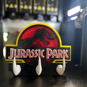 Puede incluir: Un llavero de Jurassic Park negro y amarillo con tres ganchos blancos. El logotipo de Jurassic Park presenta un fondo rojo con una silueta de dinosaurio negro y las palabras "JURASSIC PARK" en blanco.