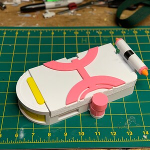 Puede incluir: Un modelo impreso en 3D blanco, rosa y amarillo de un dispositivo futurista. El dispositivo tiene una forma redondeada y presenta una serie de patrones geom&eacute;tricos. El modelo est&aacute; sentado en una estera de corte verde.