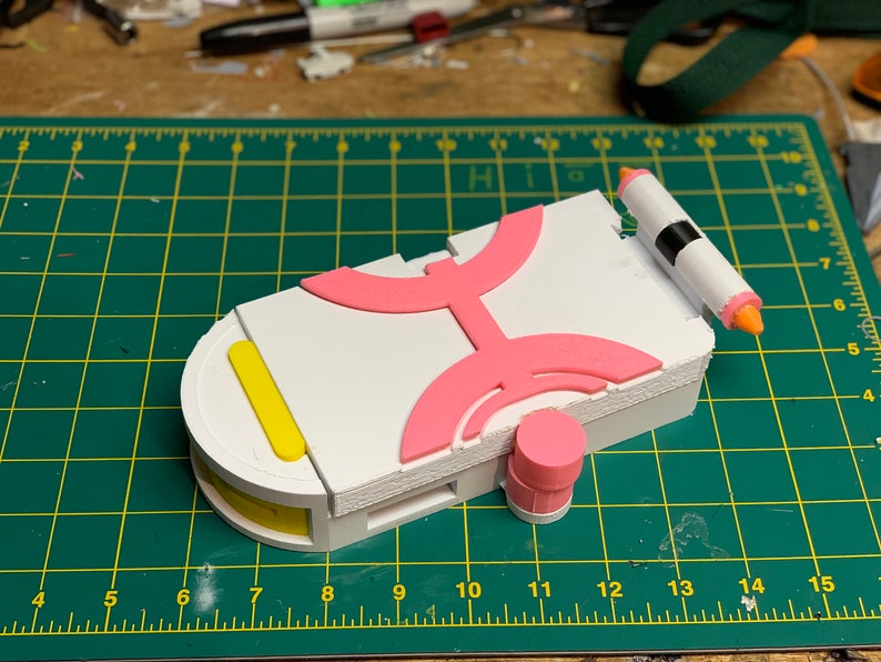 Puede incluir: Un modelo impreso en 3D blanco, rosa y amarillo de un dispositivo futurista. El dispositivo tiene una forma redondeada y presenta una serie de patrones geom&eacute;tricos. El modelo est&aacute; sentado en una estera de corte verde.
