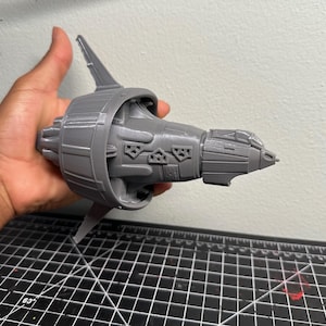 May include: A grey 3D-printed model of a spaceship engine. The model has intricate details, including fins, vents, and a cockpit-like structure. The engine is held in a hand against a plain background.