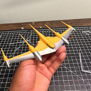Può includere: Un modello di aereo giallo e argento con tre pinne verticali e due gondole motore argentate. Il modello è tenuto in mano, mostrando il suo design e la combinazione di colori. Le ali sono argentate con dettagli gialli.