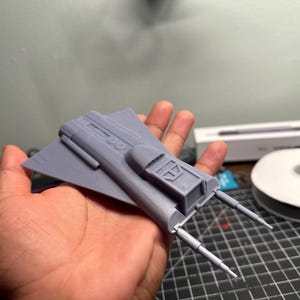 May include: A gray, 3D-printed model of a spacecraft, held in a hand. The model has a wing-like structure with a central cockpit and two forward-facing, cylindrical protrusions. The model is likely a miniature replica or a prototype.