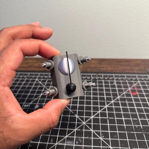 以下が含まれることがあります： 前面に円形のデザインが施された、小さなグレーの3Dプリントオブジェクト。4つの銀色の突起と、細い黒い棒に取り付けられた黒い球があります。オブジェクトは手で持たれています。