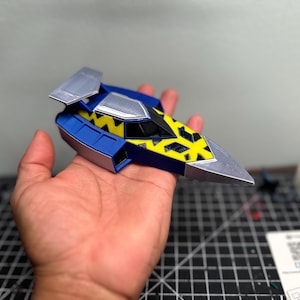 May include: A small, futuristic-looking model of a spacecraft or vehicle. The model is primarily blue with silver accents on the wings and nose. The top section features a yellow and black pattern. The model is held in a hand.