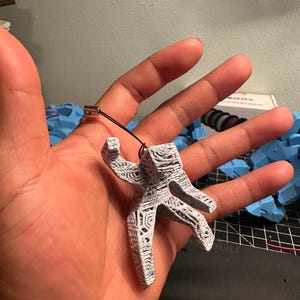 May include: A small, gray and white figurine in the shape of a person with intricate carved details. The figurine is attached to a black string with a silver clasp. The figurine is held in an open hand.