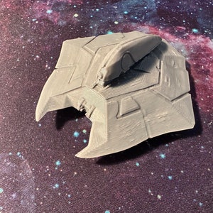May include: A gray 3D printed spaceship model with a detailed design. The model has a pointed nose and a flat bottom with a series of panels.