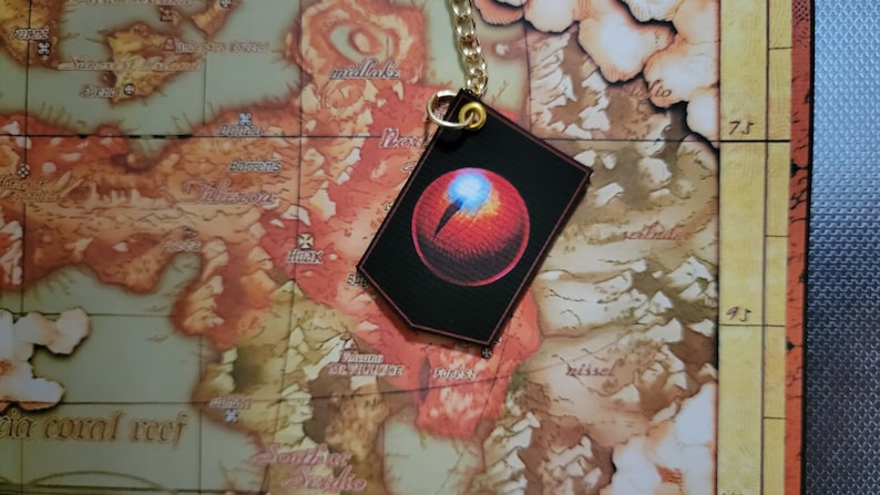 May include: A red eye with a white pupil on a black background. The eye is attached to a gold chain and is hanging from a map.