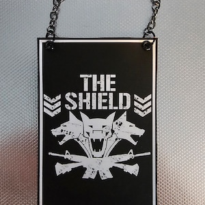 Puede incluir: Señal en blanco y negro con el texto "THE SHIELD" y un lobo de tres cabezas con rifles cruzados. La señal está colgada de una cadena.