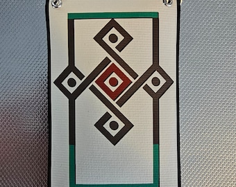 May include: A white rectangular sign with a black border and a green border inside the black border. The sign has a geometric design with black, red, and white shapes. The sign is hanging from a silver chain.