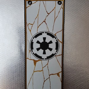 Puede incluir: Un adorno de pared rectangular con temática de Star Wars. El diseño presenta un fondo blanco con líneas doradas en forma de grietas, un borde negro y el símbolo del Imperio Galáctico en el centro. Dos remaches negros en la parte superior.