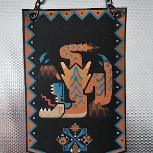 Op de afbeelding: Een zwart en bruin wandkleed met een gestileerde afbeelding van een monster met blauwe en oranje accenten. Het wandkleed heeft een ketting om op te hangen en een geometrisch patroon rond de rand.