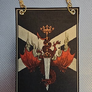 Puede incluir: Un estandarte blanco y negro con un dragón rojo enroscado alrededor de una espada plateada. El dragón lleva una corona dorada y está rodeado de humo. El estandarte tiene una cadena dorada unida a la parte superior.