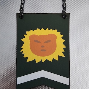 Può includere: Uno stendardo verde con una chevron bianca in basso. Un sole giallo con un viso di leone arancione è al centro. Lo stendardo ha una catena nera attaccata alla parte superiore.