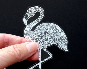 May include: A clear sticker featuring a white, detailed flamingo design. The flamingo is decorated with floral and geometric patterns. The sticker is held against a black background.