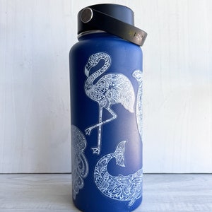 May include: A blue water bottle with a black handle and lid. The bottle is decorated with white, detailed illustrations of a flamingo, jellyfish, and whale tail. The bottle is cylindrical and appears to be made of metal.