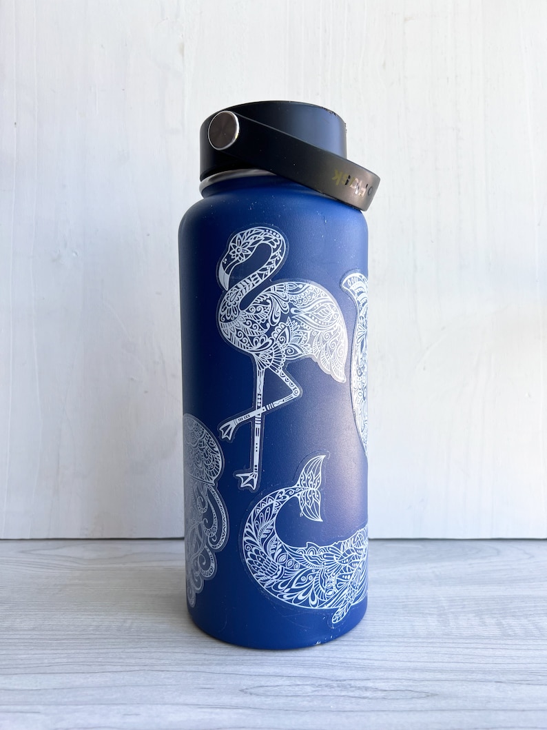 May include: A blue water bottle with a black handle and lid. The bottle is decorated with white, detailed illustrations of a flamingo, jellyfish, and whale tail. The bottle is cylindrical and appears to be made of metal.