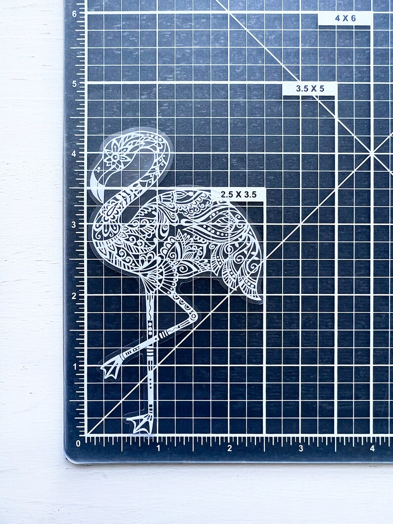 May include: A clear sticker featuring a white flamingo design. The flamingo is intricately detailed with floral and geometric patterns. The sticker measures approximately 2.5 x 3.5 inches. The flamingo is standing on one leg, with its head and neck curved.