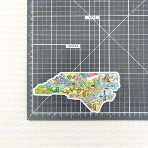 May include: A colorful sticker in the shape of North Carolina, featuring illustrations of state landmarks and icons. The sticker measures 3.5 x 5 inches and includes the text "Tar Heel" and "First Flight".