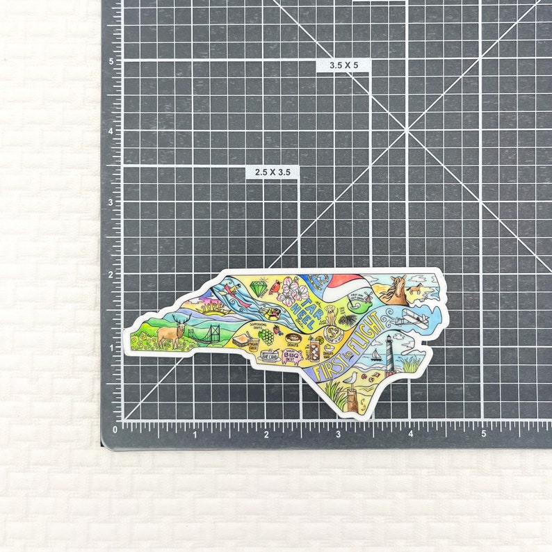 May include: A colorful sticker in the shape of North Carolina, featuring illustrations of state landmarks and icons. The sticker measures 3.5 x 5 inches and includes the text "Tar Heel" and "First Flight".