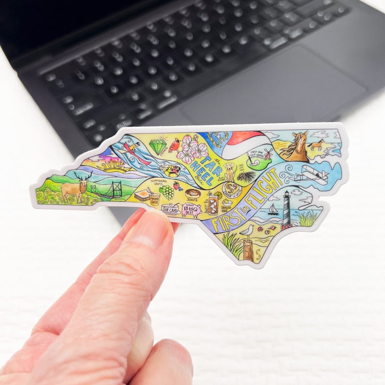 May include: A colorful sticker in the shape of North Carolina, featuring illustrations of state landmarks and icons. The sticker includes the text "TAR HEEL" and "FIRST FLIGHT". The design incorporates a deer, a lighthouse, and a horse.