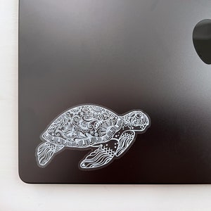 May include: A white sticker of a detailed, ornate sea turtle with a floral pattern on a black laptop.