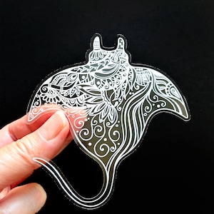May include: White vinyl sticker of a stylized manta ray with intricate patterns and swirls. The sticker is on a black background.