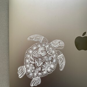 May include: A white vinyl sticker of a detailed, ornate, stylized turtle with a floral design on a silver laptop.