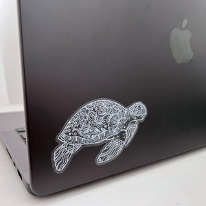May include: A white sticker of a detailed, ornate turtle design is placed on the back of a silver laptop.