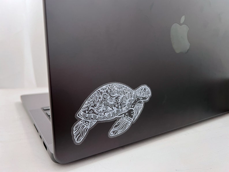 May include: A white sticker of a detailed, ornate turtle design is placed on the back of a silver laptop.