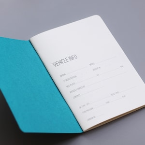 Puede incluir: Una libreta blanca con una cubierta turquesa, abierta en una página con el encabezado "VEHICLE INFO". La página tiene secciones para "Marca", "Registro", "Placa", "Propietario anterior", "Contacto", "Modelo", "Comprado en", y "Kilometraje".