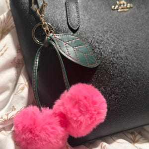 May include: A black leather handbag with a gold-colored Coach logo and a pink fluffy cherry keychain. The keychain has a green leaf with red stitching and a gold clasp.