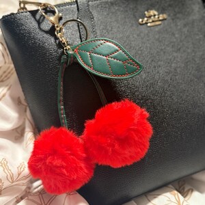 May include: A black leather handbag with a gold zipper and a cherry-shaped keychain. The keychain features two fluffy red pom-poms and a green leaf with red stitching. The bag has a gold Coach logo.