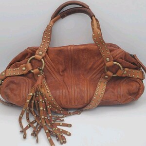 May include: A brown leather handbag with a distressed finish. The bag features studded straps, a tassel, and metal ring accents. The handles are a darker brown. The bag is approximately 14 inches wide.