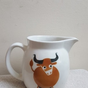 May include: White ceramic pitcher with a brown bull design. The bull has black horns and eyes.