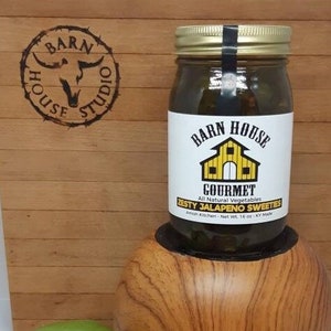 May include: A glass jar of Zesty Jalapeno Sweeties, a gourmet, all-natural vegetable product from Barn House Amish Kitchen. The jar is sitting on a wooden surface with two tomatoes.