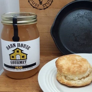 May include: A glass jar of pear preserves with a yellow and brown label that reads "Barn House Gourmet All Natural Preserves Pear Amish Kitchen - Net Wt 19 oz - KY Made". Two golden brown biscuits are on a white plate next to a black cast iron skillet.