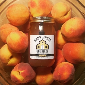 May include: A glass jar of peach preserves with a label that reads "Barn House Gourmet All Natural Preserves Peach Amish Kitchen - Net Wt. 19 oz. KY Made". The jar is surrounded by fresh peaches.
