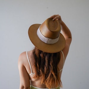 Puede incluir: Un sombrero fedora marrón con una banda de patrón blanco y gris. El sombrero está hecho de paja y tiene un ala ancha.
