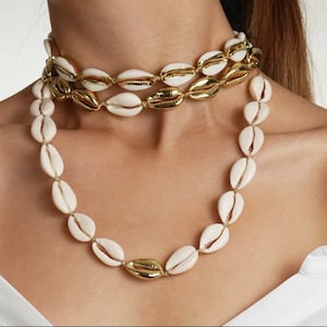 Puede incluir: Un collar de capas hecho de conchas blancas y detalles dorados. El collar es de estilo gargantilla y tiene una hebra más larga de conchas que cuelga en la parte delantera.