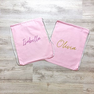 May include: Two pink drawstring bags with the names "Isabella" and "Olivia" embroidered on them in purple and gold glitter.