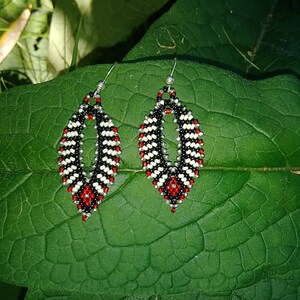 May include: A pair of dangle earrings with a black, white, and red beaded design. The earrings are shaped like teardrops and have a silver hook.