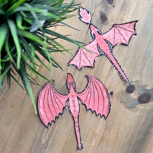 May include: Two pink paper dragons with black outlines and white details. The dragons are cut out and have a whimsical, fairytale-like style.