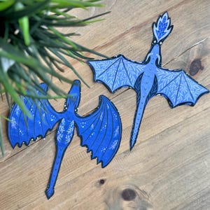 May include: Two blue dragon cutouts with intricate wing and body designs. The dragons have a stylized appearance with detailed patterns and a celestial theme. The wings are spread, and the dragons are positioned on a wooden surface.