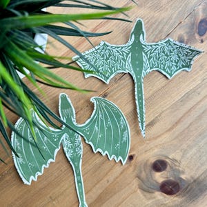 May include: Two green paper dragon cutouts with a leafy pattern. The dragons are facing opposite directions with their wings spread out.